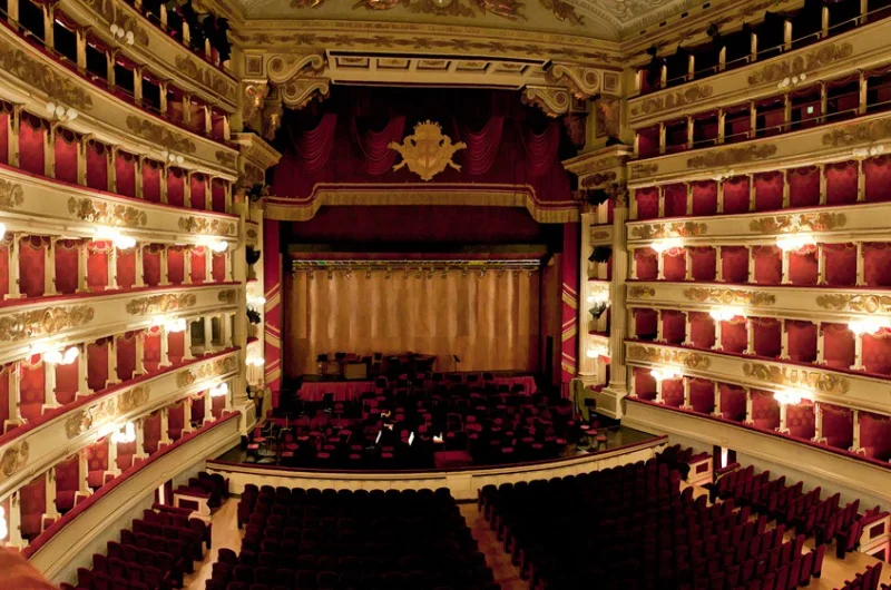 Widok wnętrza Teatro alla Scala w Mediolanie, ukazujący zabytkowy gmach opery z czerwonymi aksamitnymi balkonami, ozdobnymi dekoracjami i główną sceną.