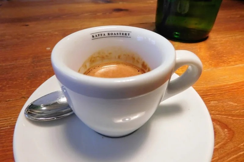 Eszpresszó fehér csészében és tányéron, egy Kaffa Roastery logóval ellátott faasztalon, a csésze mellé egy kanállal tálalva.