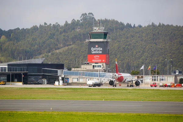 A Coruña Airport control tower and terminal buildings with an Iberia Express aircraft parked on the tarmac, surrounded by forested hills and green grass areas.