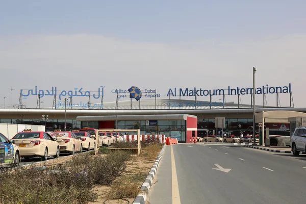 Al Maktoum International Airport entrance with signage, parking area, and vehicles on the road leading to the terminal building.