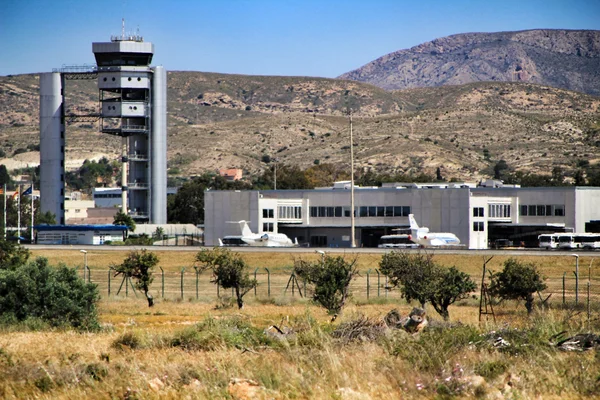 Air traffic control tower and terminal buildings at Alicante Elche Miguel Hernández Airport surrounded by arid hillsides and olive trees.