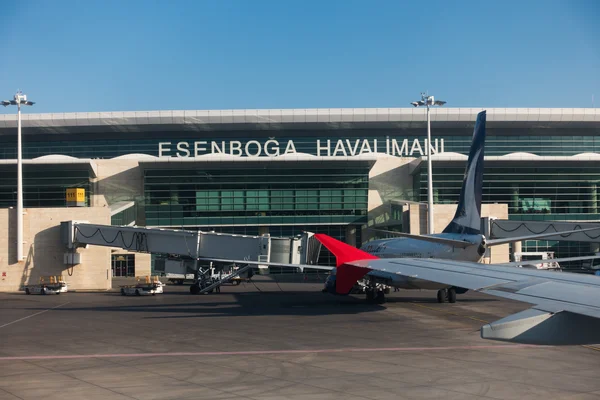 Ankara Esenboğa Airport terminal building with aircraft wing in foreground and ground support equipment visible under clear blue sky.