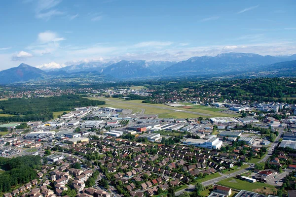 Vista aérea del Aeródromo de Annemasse con áreas residenciales en primer plano y montañas alpinas visibles bajo cielo despejado.
