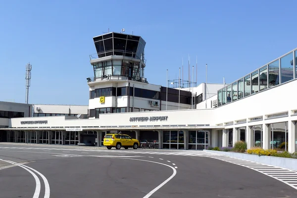Terminal del Aeropuerto Internacional de Amberes con torre de control, vehículo amarillo y arquitectura moderna bajo cielo azul despejado.