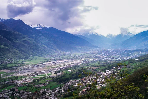 Aerial view of Aosta Valley Airport nestled in an alpine valley surrounded by snow-capped mountains and green landscapes.