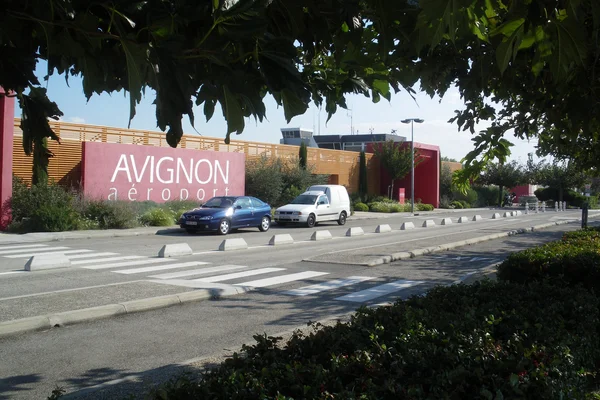 Entrada del Aeropuerto de Aviñón Provenza con señalización moderna, zona de estacionamiento y vehículos bajo sombra de árboles en día soleado.