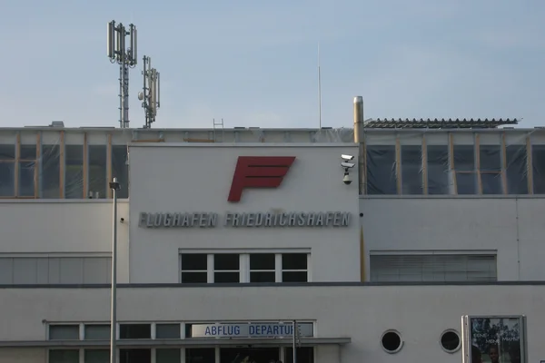 Edificio terminal del Aeropuerto Bodensee Friedrichshafen con logo rojo 'F', antenas de comunicación en techo y señalización de aeropuerto en fachada blanca.