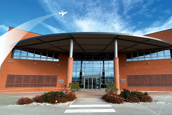 Modern terminal building at Bologna Guglielmo Marconi Airport with red brick facade, large windows, and aircraft flying overhead against blue sky.