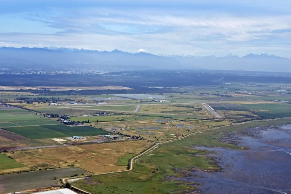 Vue aérienne de l'aéroport de Boundary Bay avec terres agricoles, zones humides et montagnes sous un ciel partiellement nuageux.