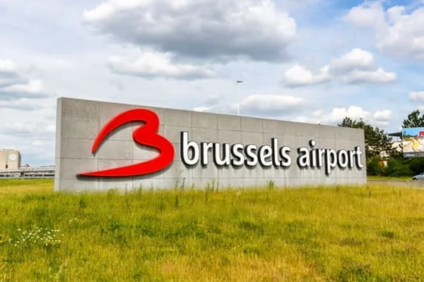 Gran letrero del Aeropuerto de Bruselas con logo rojo en campo herboso bajo cielo parcial.