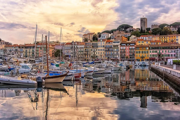 Coloridos edificios frente al agua y veleros reflejados en aguas tranquilas del puerto al atardecer en Cannes, Francia.