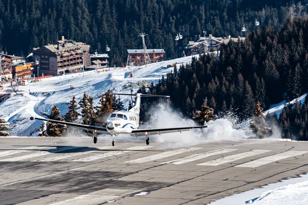 Mały samolot ląduje na zaśnieżonym pasie startowym w Courchevel Altiport, otoczony alpejskim lasem i stokami narciarskimi z widocznymi budynkami górskimi w tle.