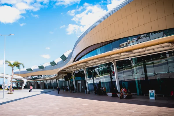 Modern curved architecture of Faro Airport terminal with bronze paneling, large windows, and a spacious plaza under blue skies.
