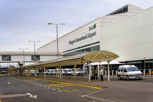 Wejście do terminalu Glasgow International Airport ze strefą załadunku, białymi pojazdami i nowoczesną fasadą budynku pod pochmurnym niebem.