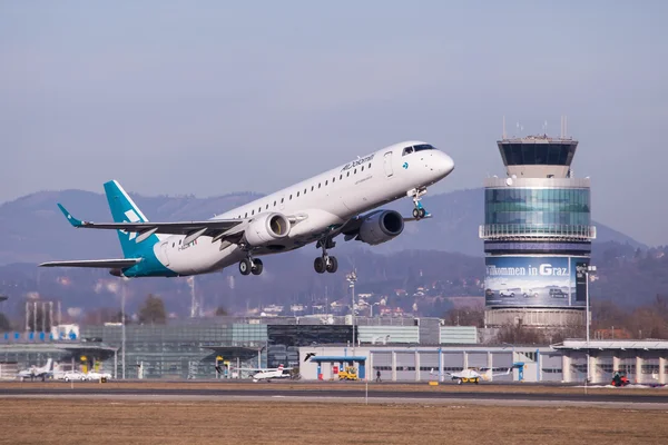 Aeronave comercial azul verdosa y blanca despega del Aeropuerto de Graz con torre de control visible al fondo.