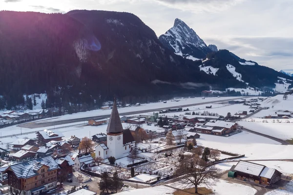 Widok z lotu ptaka lotniska Saanen Gstaad w zaśnieżonej alpejskiej dolinie z wieżą kościoła na pierwszym planie i ośnieżonymi górami w tle.