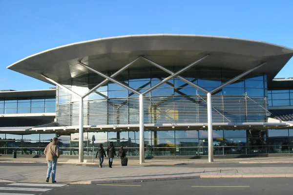 Edificio terminal moderno del Aeropuerto de Guernsey con techo metálico curvo y fachada de vidrio, viajeros afuera en día despejado.