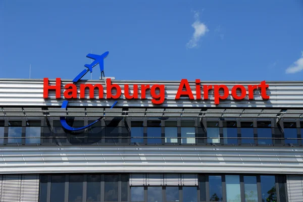 Exterior del edificio terminal del Aeropuerto de Hamburgo con señalización roja y símbolo azul de avión contra cielo azul despejado.