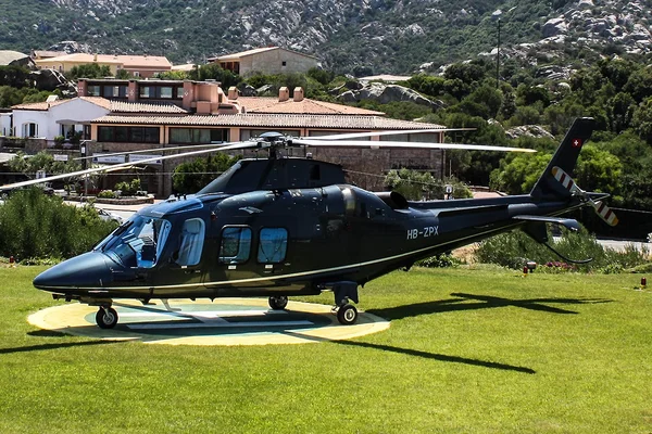 Black helicopter HB-ZPM parked on green grass at Helipad Villa La Contra with mountainous terrain and buildings in the background.