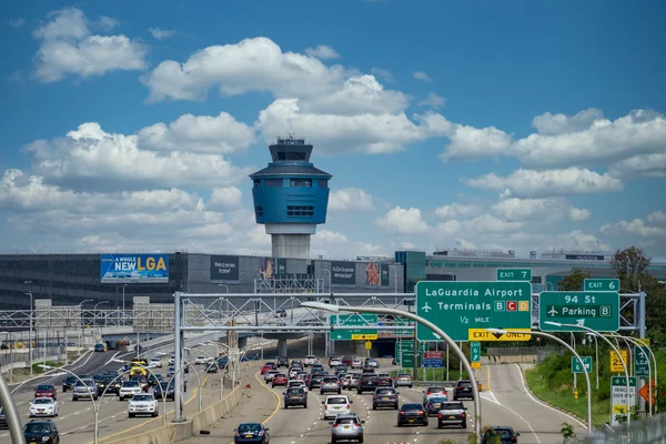 Tour de contrôle et terminal de l'aéroport LaGuardia avec trafic routier et ciel bleu nuageux visible.