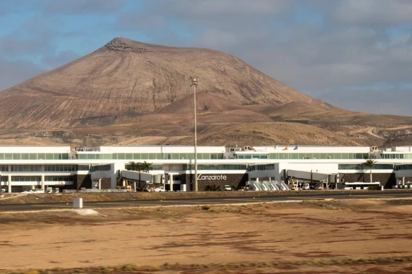 Modern airport terminal building with white and green exterior stands before a distinctive cone-shaped volcanic mountain under a partly cloudy sky.