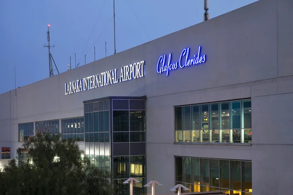 Larnaca International Airport terminal building exterior at dusk, displaying illuminated signage on the modern gray facade with large windows and communication antennas on the roof.