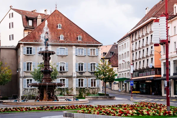 Historyczny europejski rynek z centralną fontanną, kolorowymi rabatami kwiatowymi i tradycyjną architekturą z czerwonymi dachami otaczającymi plac.