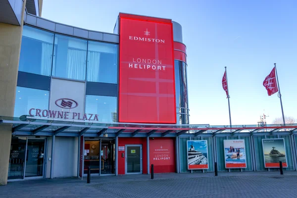 Wejście do budynku terminalu London Heliport z czerwonym znakiem, szklannymi oknami i widocznym po lewej stronie hotelem Crowne Plaza.
