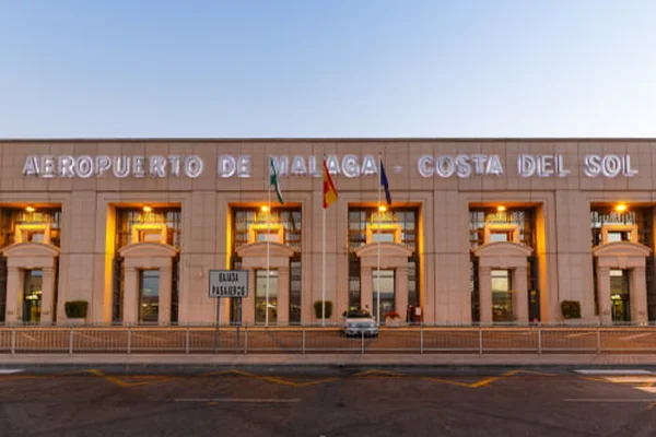 Exterior facade of Málaga Costa del Sol Airport terminal building with illuminated arched windows and Spanish flags at dusk.