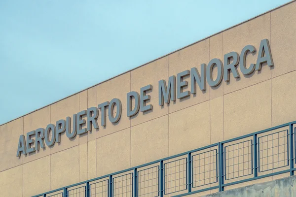 Exterior view of Menorca Airport terminal building with blue lettering on tan facade and blue metal fencing below.