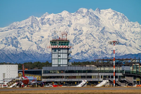Milan Malpensa Airport control tower with snow-capped Alpine mountains in the background under clear blue sky.