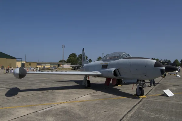 A military jet aircraft parked on the tarmac at Monte Real Air Base with support vehicles and hangars visible in the background.