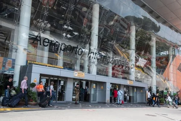 Exterior view of Naples International Airport terminal building with glass facade, white columns, and passengers entering the entrance area.
