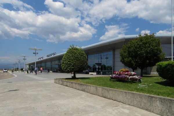Modern terminal building of Podgorica Airport with landscaped gardens, flowering plants, and clear sky with clouds overhead.