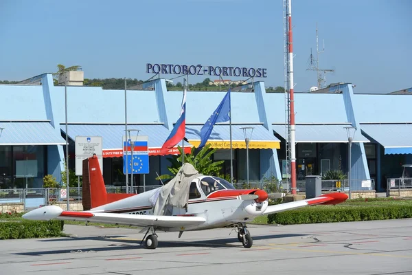 A small white and red aircraft parked in front of the Portorož Airport terminal building with flags displayed.