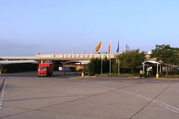 Exterior view of Reus Airport terminal building with red vehicle parked outside and Spanish flags visible on a clear day.
