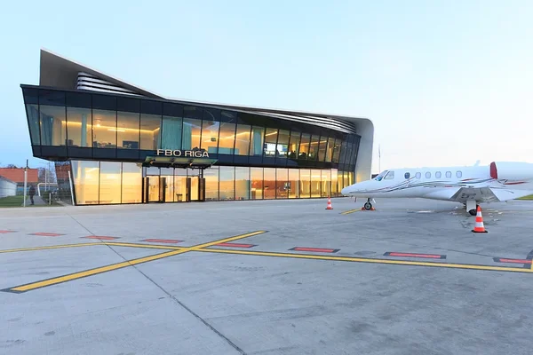 Terminal FBO moderne de Riga avec façade en verre illuminée au crépuscule et jet privé garé.