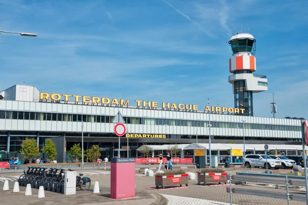 Budynek terminalu Lotniska Rotterdam The Hague z czerwono-białą wieżą kontroli ruchu lotniczego pod błękitnym niebem.
