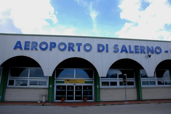 Exterior view of Salerno Costa d'Amalfi Airport terminal building with blue signage, green doors, and distinctive arched windows against a cloudy sky.