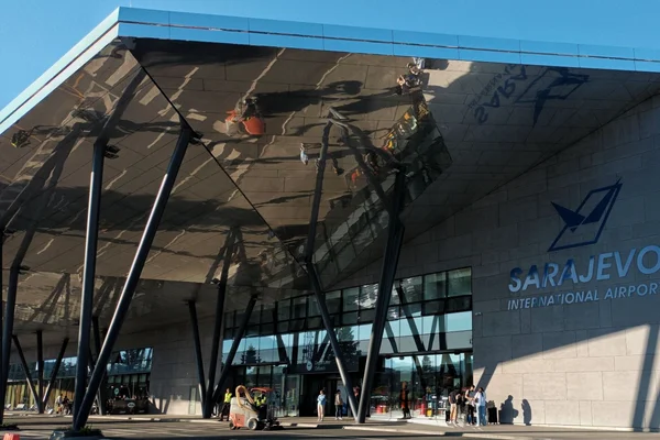Sarajevo International Airport terminal entrance with modern architecture, distinctive black steel canopy structure, and blue signage on a sunny day.