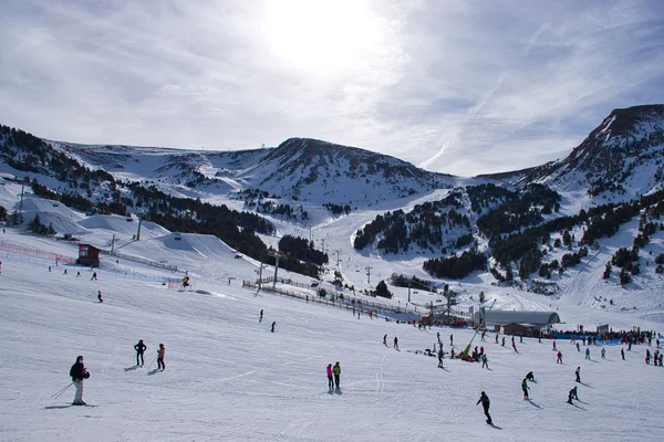 A snowy ski resort nestled in mountain valleys with skiers on the slopes and ski lifts visible under a partly cloudy sky.