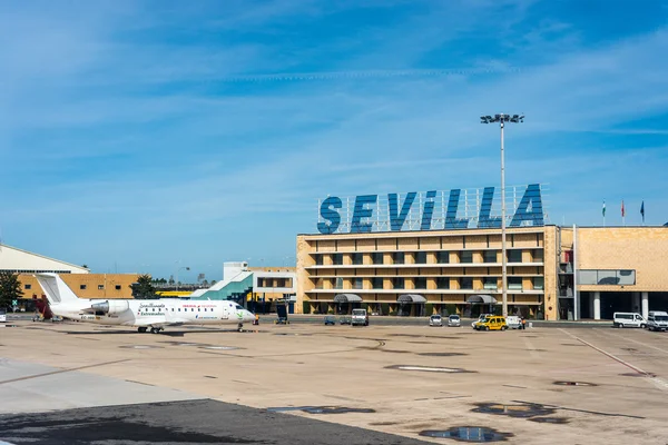 Seville Airport terminal building with large blue lettering on a sunny day, featuring a parked aircraft and ground vehicles on the tarmac.