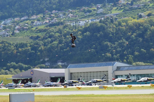 Paralotniarze schodzący nad lotniskiem Sion z widocznymi hangarami i zaparkowanymi samolotami, na tle zalesionych Alp i budynków mieszkalnych.