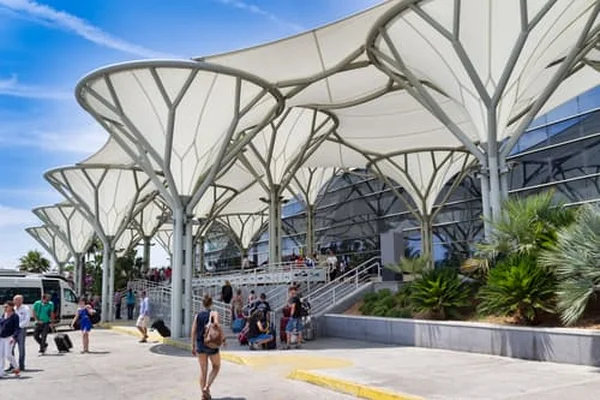Modern airport terminal with distinctive white petal-shaped canopy structures, palm trees, and travelers walking in the sunny plaza area at Split Saint Jerome Airport.