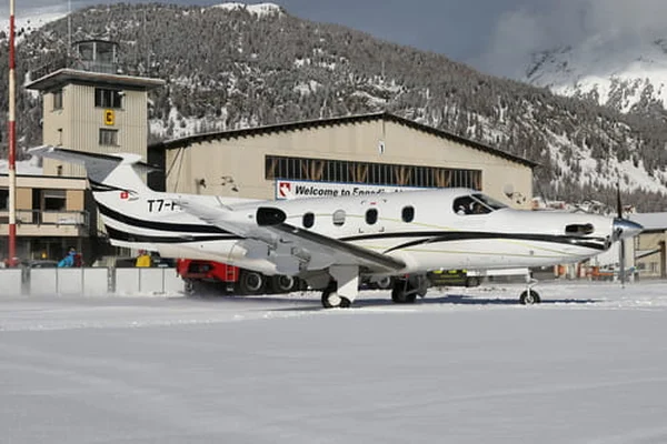 Dwusilnikowy samolot zaparkowany na zaśnieżonym asfalcie na lotnisku Samedan, ze śnieżnymi górami i budynkiem hangaru widocznym w tle.