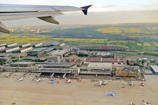 Widok z lotu ptaka na lotnisko w Stuttgarcie z skrzydłem samolotu na pierwszym planie, budynkami terminala, zaparkowanymi samolotami i zieloną okolicą w tle.