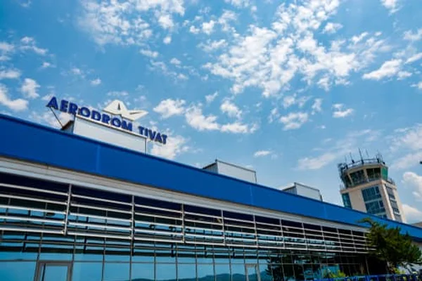 Exterior view of Tivat Airport terminal building with blue signage, control tower, and clear sky with clouds.