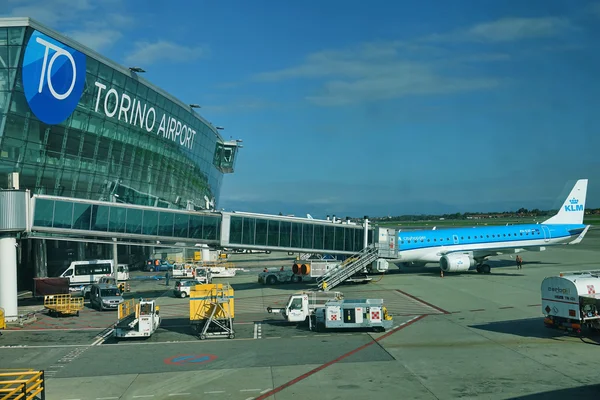 A blue KLM aircraft parked at Turin Airport's Terminal 10, with ground service vehicles and equipment visible on the tarmac under clear skies.