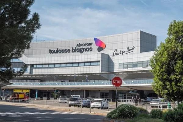 Terminal Lotniska Toulouse-Blagnac z nowoczesną fasadą ze szkła, kolorowym logo i zaparkowanymi pojazdami na przedzie pod jasnym niebem.