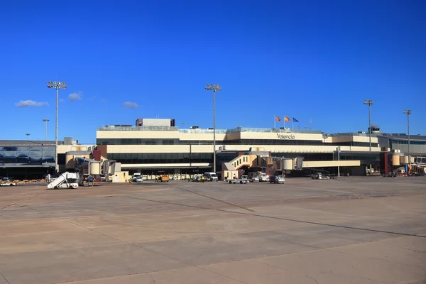 A large modern airport terminal building at Valencia Airport under clear blue skies, with aircraft service vehicles and ground equipment visible on the tarmac.